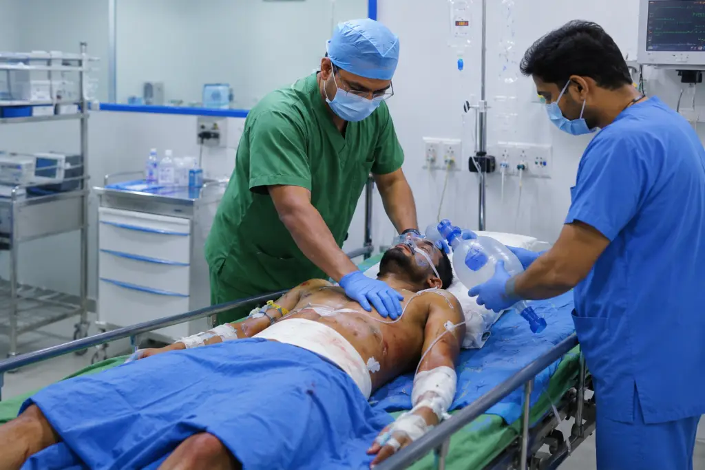 Two clinicians in scrubs attend a male patient on a hospital gurney; one checks his chest while the other provides ventilation with a bag‑valve mask and oxygen mask on his face, in an urgent care setting.