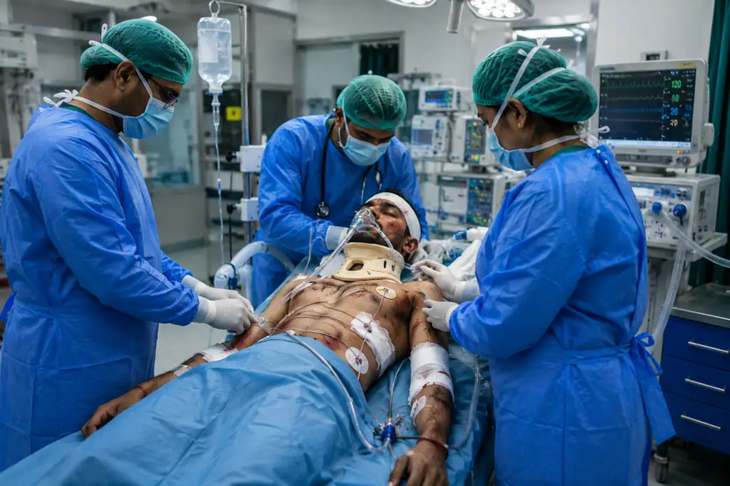 Medical team in blue gowns and masks administers care to a patient on an operating table with a neck brace and oxygen mask. A monitor and IV drip are visible in the background.