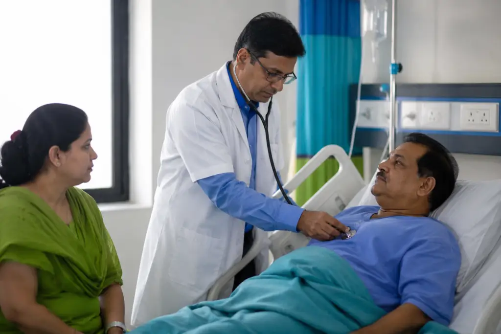 Doctor using stethoscope to listen to a patient in bed, with a woman caregiver seated beside the bed in a hospital room.