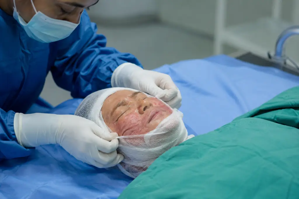 Surgeon in blue scrubs applying a bandage to a patient's head in an operating room.