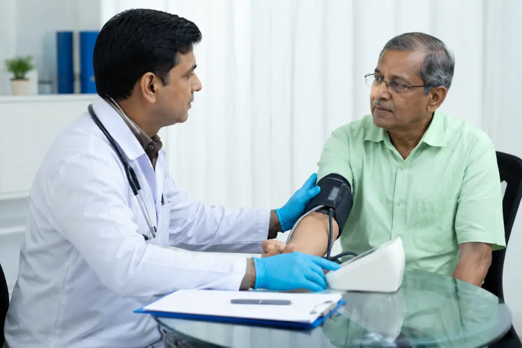Doctor measuring a patient's blood pressure with a cuff on the upper arm in a clinical setting, both seated at a glass table.