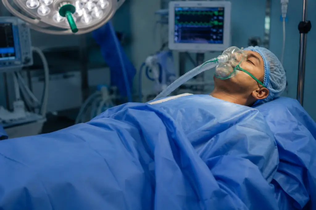 Patient on an operating table with blue surgical drapes, wearing an anesthesia mask connected to tubing. Monitors in background display vital signs.