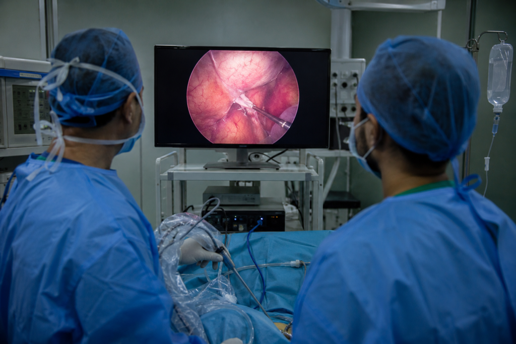 Two surgeons in blue gowns monitor a laparoscopic procedure, viewing a monitor showing internal pink tissue and instruments.