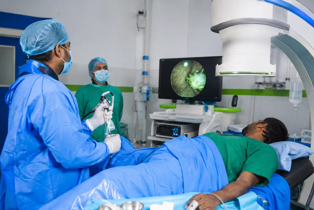 Surgeon in blue scrubs and mask performing an endoscopy while a patient lies on the operating table with a monitor showing the internal view.