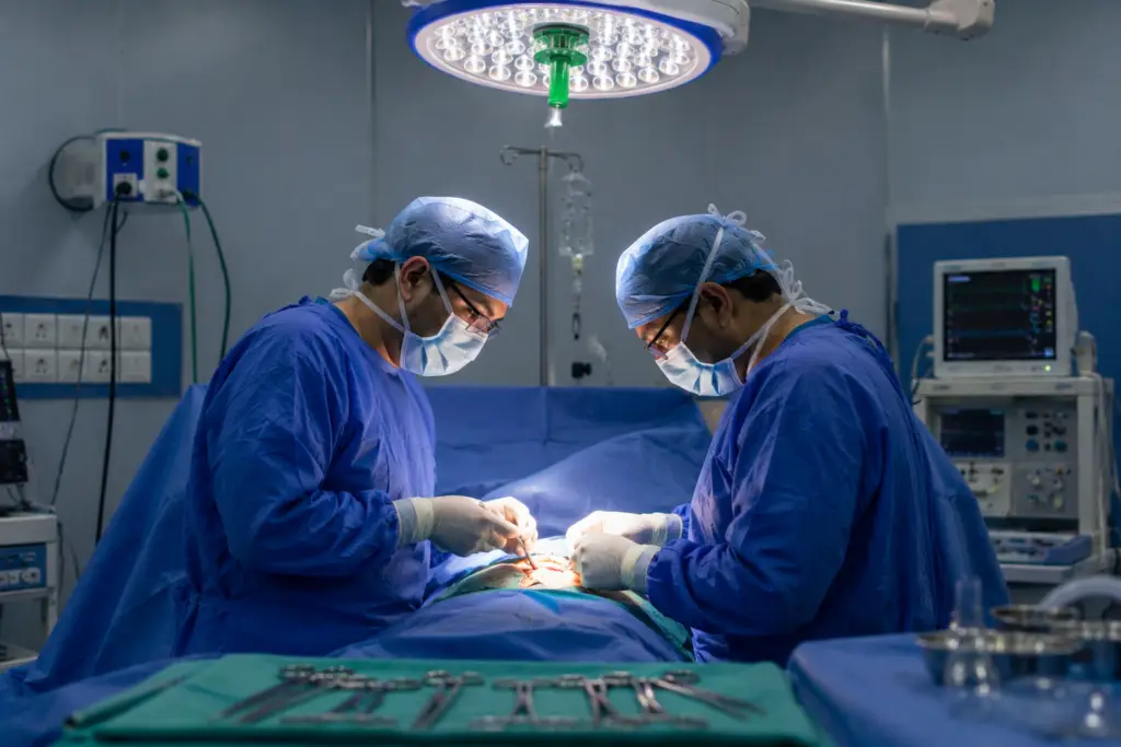 Two surgeons in blue gowns and masks perform surgery under bright overhead lights in an operating room, focused on the patient."