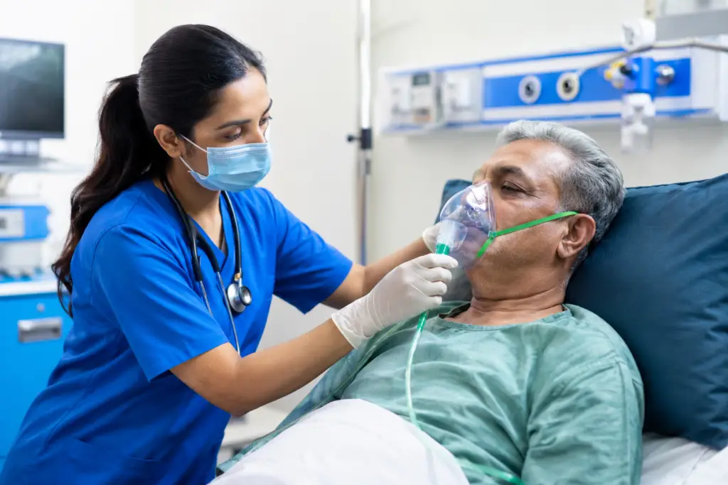 Nurse in blue scrubs, mask, and gloves administers oxygen to an elderly man wearing a clear oxygen mask in a hospital bed.