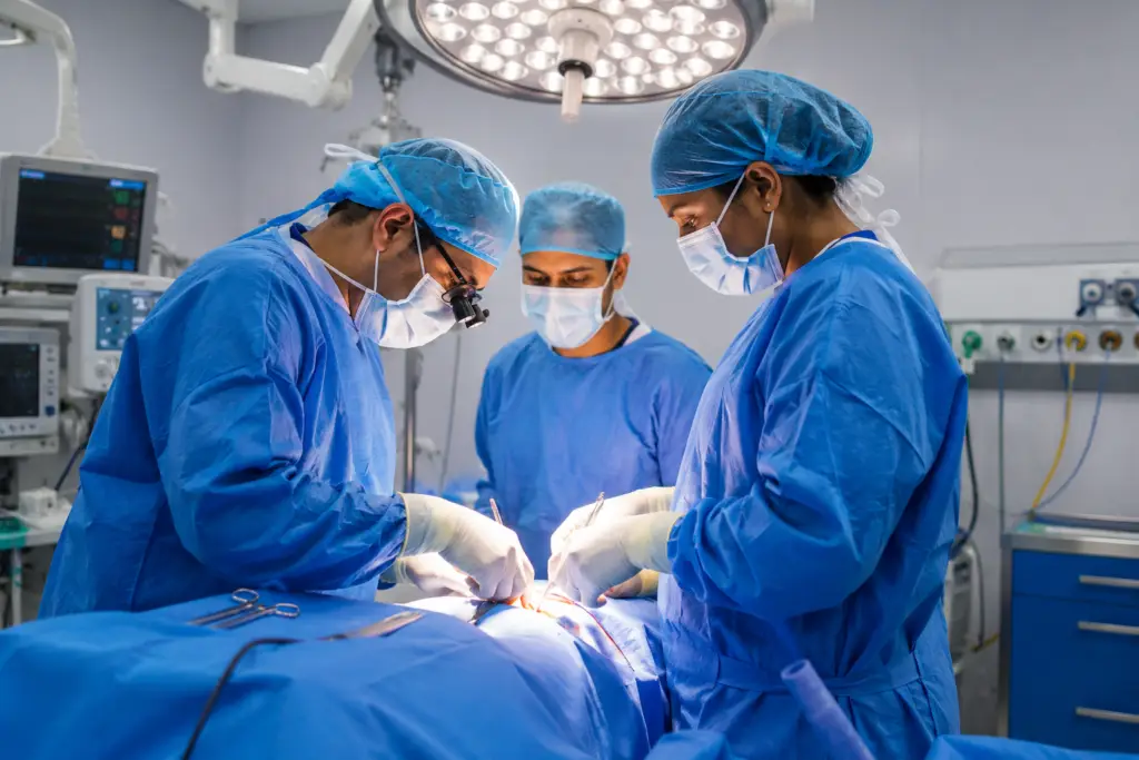 Three surgeons in blue scrubs and masks perform surgery under a bright overhead light.